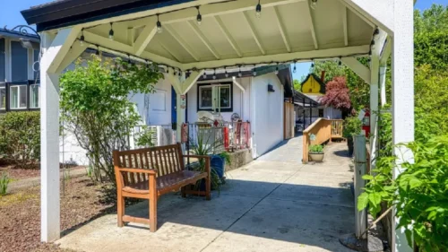 A covered patio with string lights, a wooden bench, and a paved walkway leading to a building