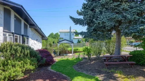 A grassy area with a dirt path, a picnic table, and a gazebo, with the side of a building on the left