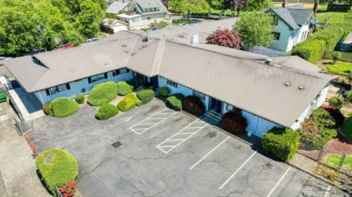 An aerial view of a building with a gray roof and a parking lot, surrounded by green hedges and trees